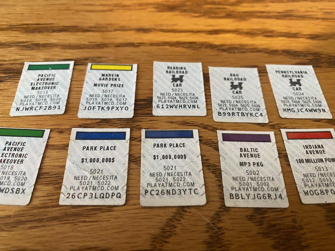 Nine Monopoly game pieces from McDonald's are arranged in two rows on a wooden surface. Each piece displays a color bar, property name, prize, and a unique code.