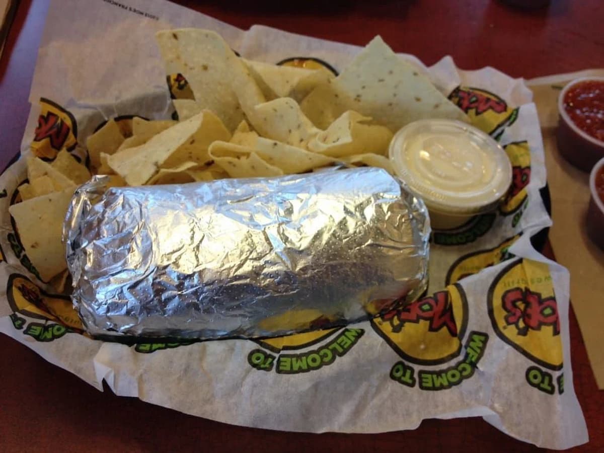 A foil-wrapped burrito served in a basket with tortilla chips, a cup of white dipping sauce, and branded paper liner—fast food done right. Part of a salsa container is visible on the side, just as you'd expect from fast-food chains that never use a microwave.