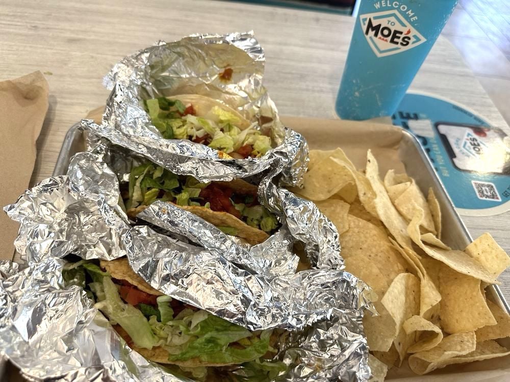 Two foil-wrapped tacos filled with lettuce, tomato, and cheese on a tray, served with tortilla chips. A blue "Welcome to Moe's" cup is in the background on a light wooden table.