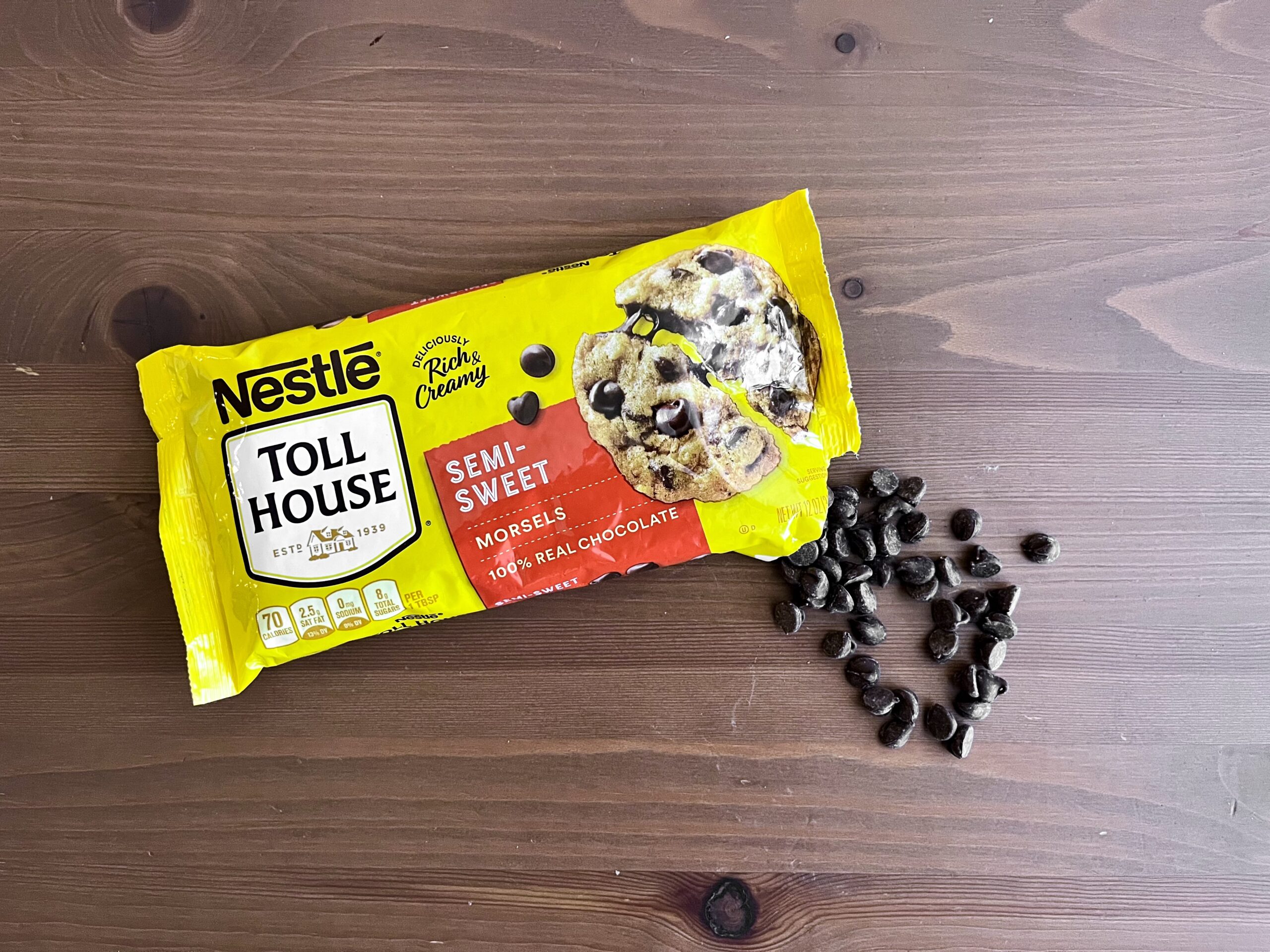 A yellow package of Nestlé Toll House semi-sweet chocolate morsels on a wooden surface, with some chocolate chips spilled next to the open bag.