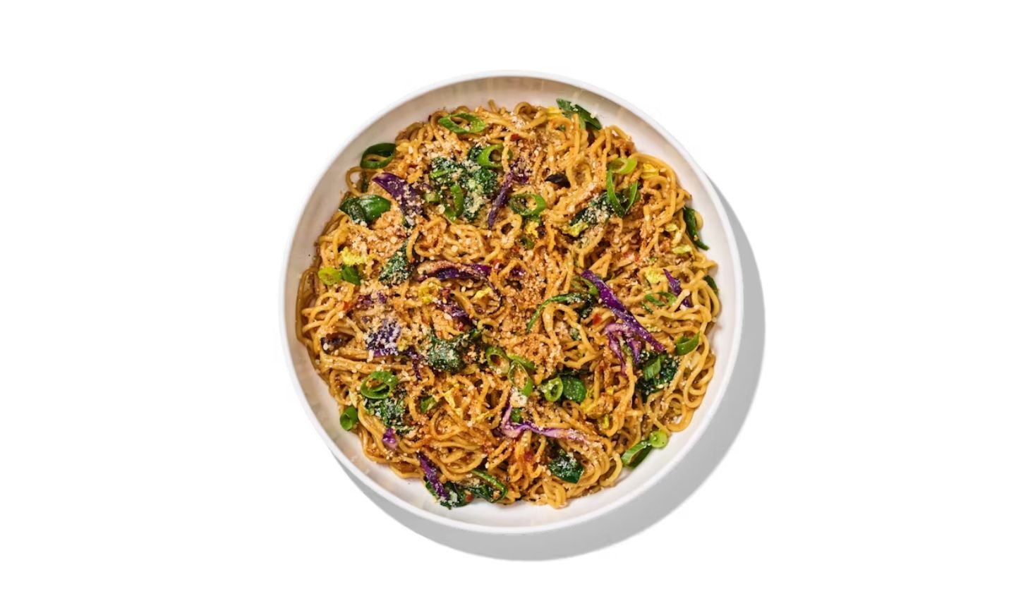 A white bowl filled with seasoned noodles, green vegetables, and purple cabbage, garnished with chopped herbs and sesame seeds—a colorful option from a new fast-food menu, presented on a clean white background.