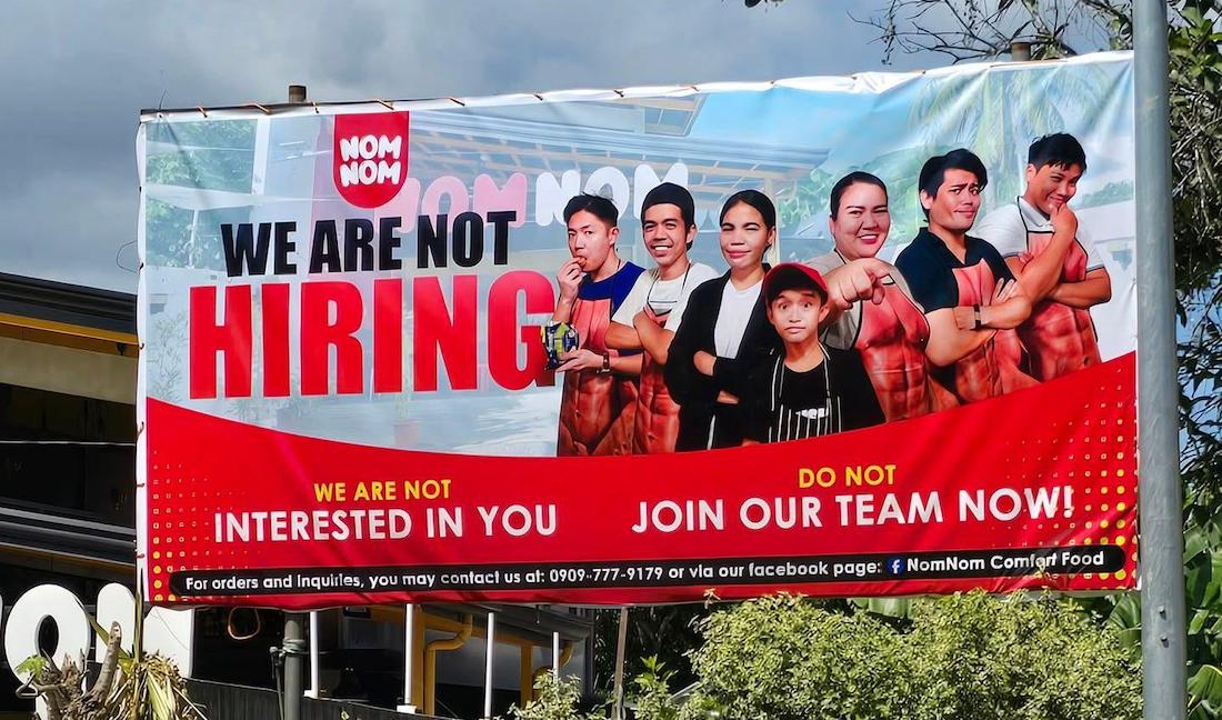 A large outdoor banner shows six adults and one child posing confidently. It reads, "WE ARE NOT HIRING. WE ARE NOT INTERESTED IN YOU. DO NOT JOIN OUR TEAM NOW!" with NomNom Comfort Food branding.