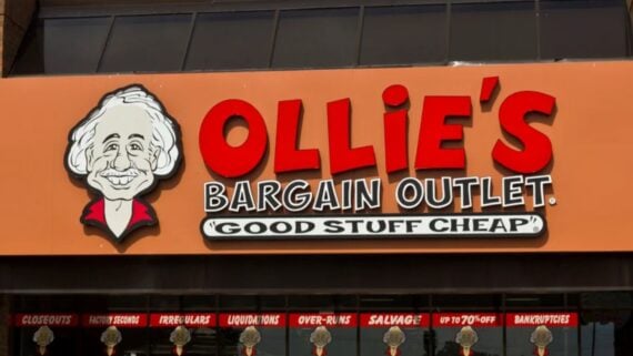 Storefront sign for Ollie's Bargain Outlet featuring a cartoon man's face and slogan "Good Stuff Cheap." The sign is orange with bold red and white lettering.