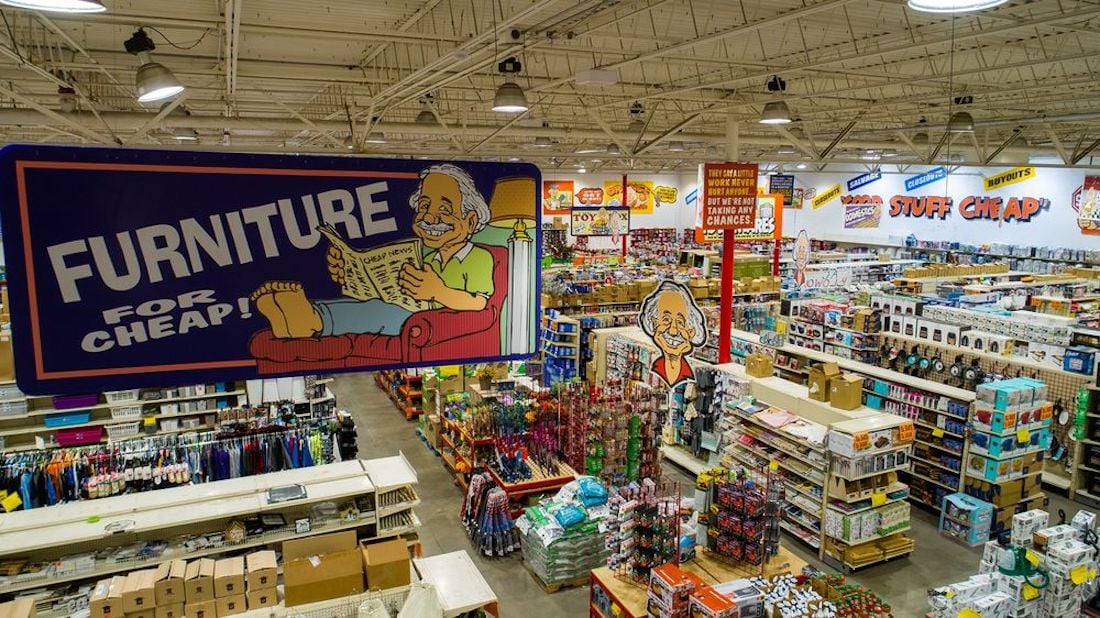 A large discount store interior with wide aisles, filled shelves, and colorful signs. A big sign reads “Furniture for Cheap!” with a cartoon man on a couch. More products and sale signs are visible throughout the store.