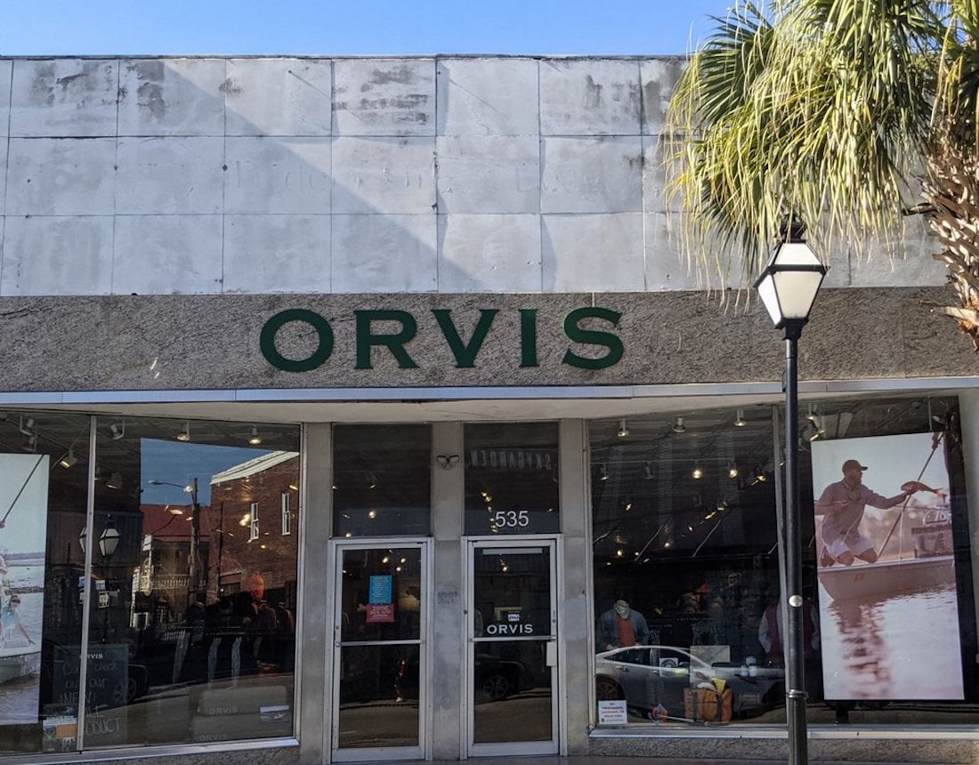A storefront with large glass windows displays the name "Orvis" above the entrance. A palm tree and streetlamp are visible outside, and fishing gear and mannequins can be seen inside.