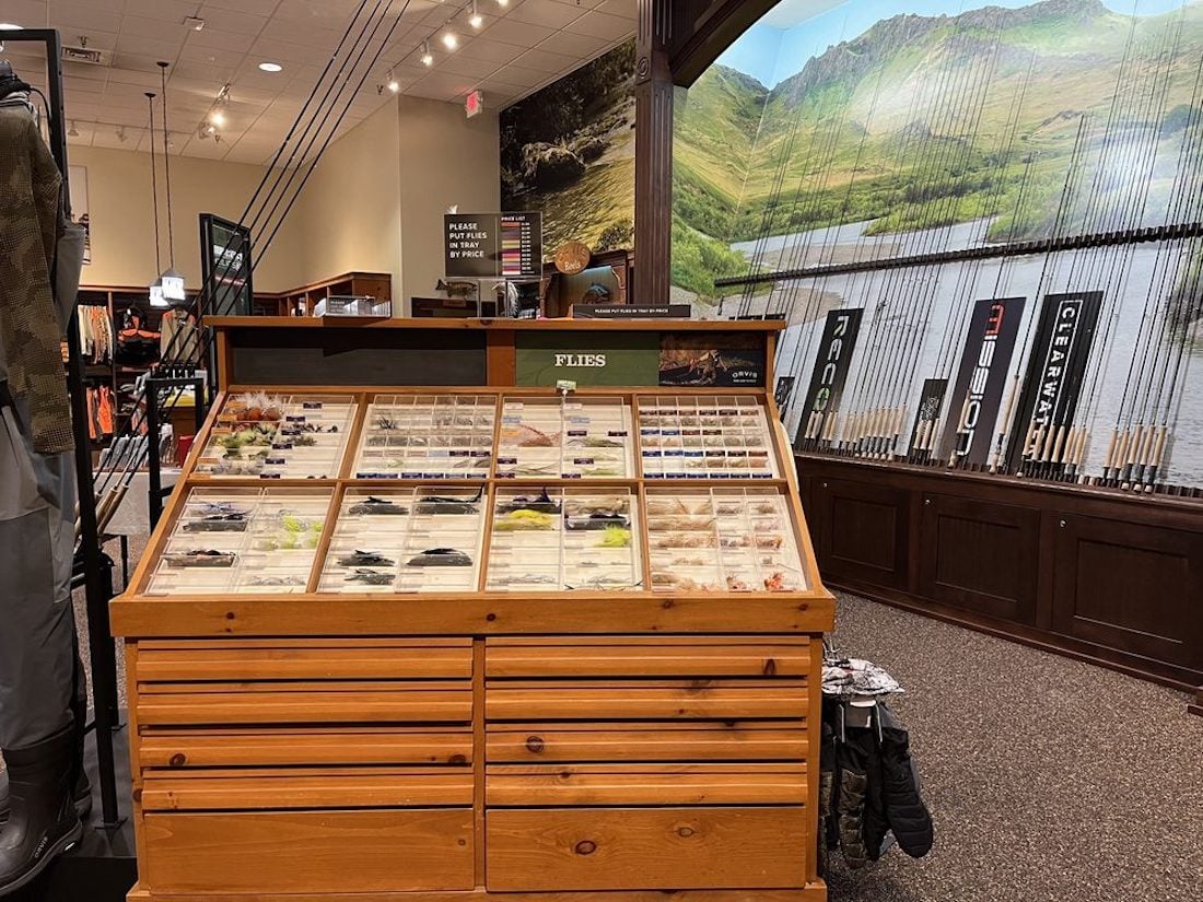 A wooden display case filled with various fishing flies is in the center of an outdoor store, with fishing rods lined up along the wall and a nature mural of mountains and a lake in the background.