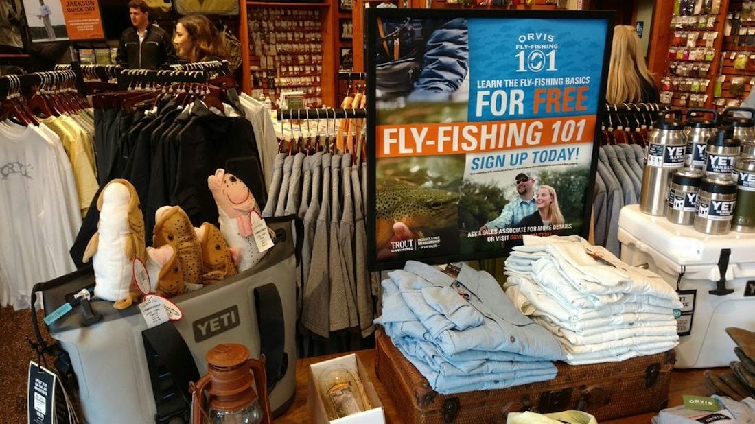 A display inside an outdoor store features shirts, Yeti coolers and tumblers, plush fish toys, and a large sign advertising free Fly-Fishing 101 classes with an image of a couple fishing.