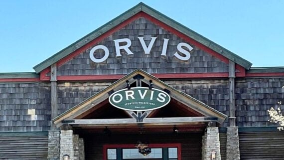 A rustic building with a shingled facade and stone columns displays two signs reading "Orvis." One sign is large and white above, and a green oval sign with "Orvis Hunting Traditions" hangs below.
