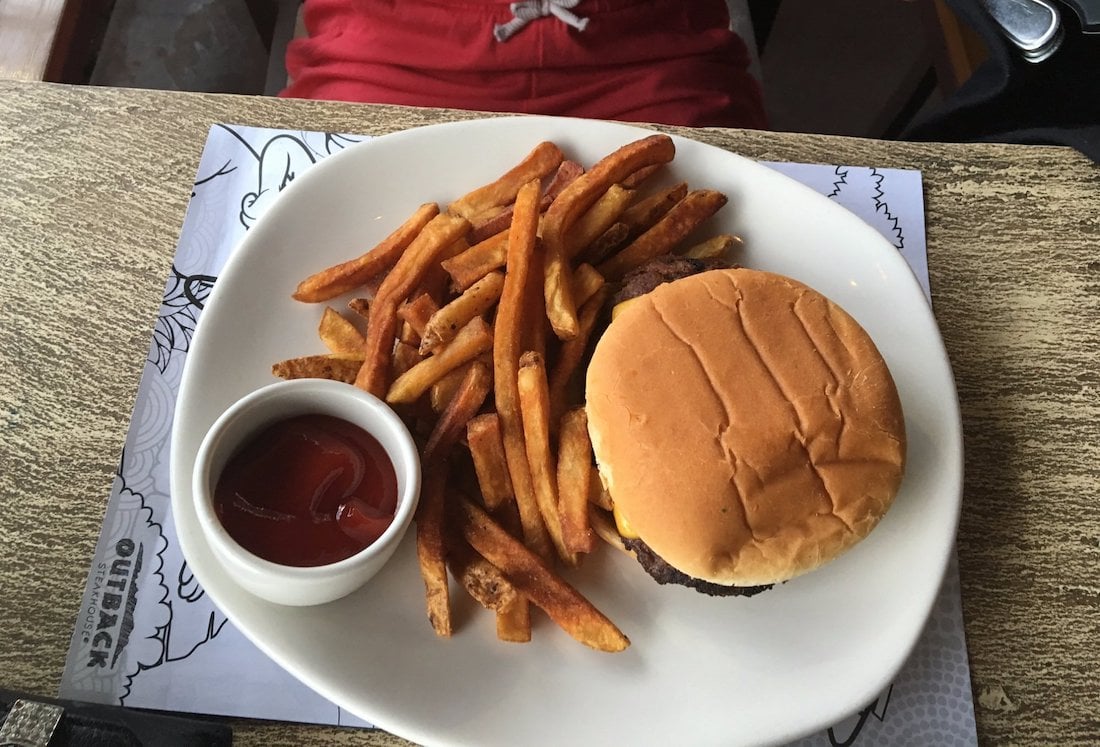 A plate with a hamburger, a pile of French fries, and a small cup of ketchup sits on a wooden table. A person wearing red shorts is partially visible in the background.