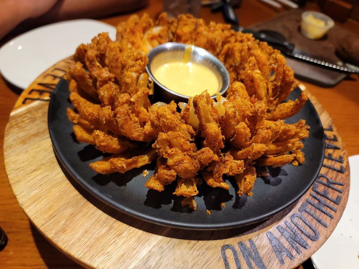 A crispy, golden-brown blooming onion is served on a black plate with a side of creamy dipping sauce in the center, set on a round wooden serving board.