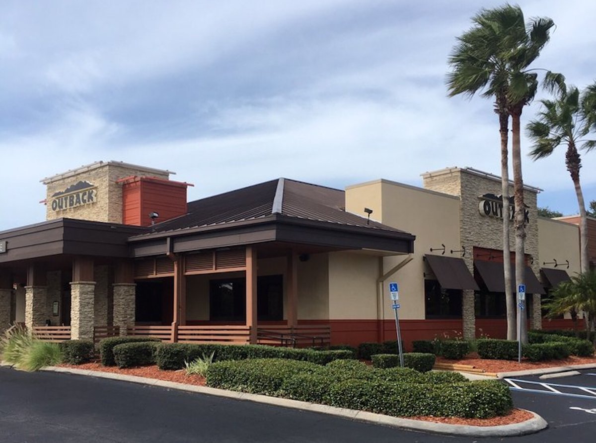 A single-story Outback Steakhouse restaurant with stone and tan exterior, brown roof, and covered patio. Palm trees and landscaped bushes surround the building under a partly cloudy sky.
