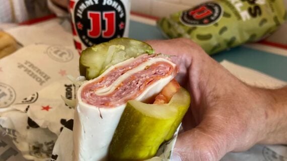 A hand holds an unwrapped Jimmy John’s sub made with layers of deli meat and cheese instead of bread, with large pickle slices as the outer layer. A branded drink cup and bag are in the background.