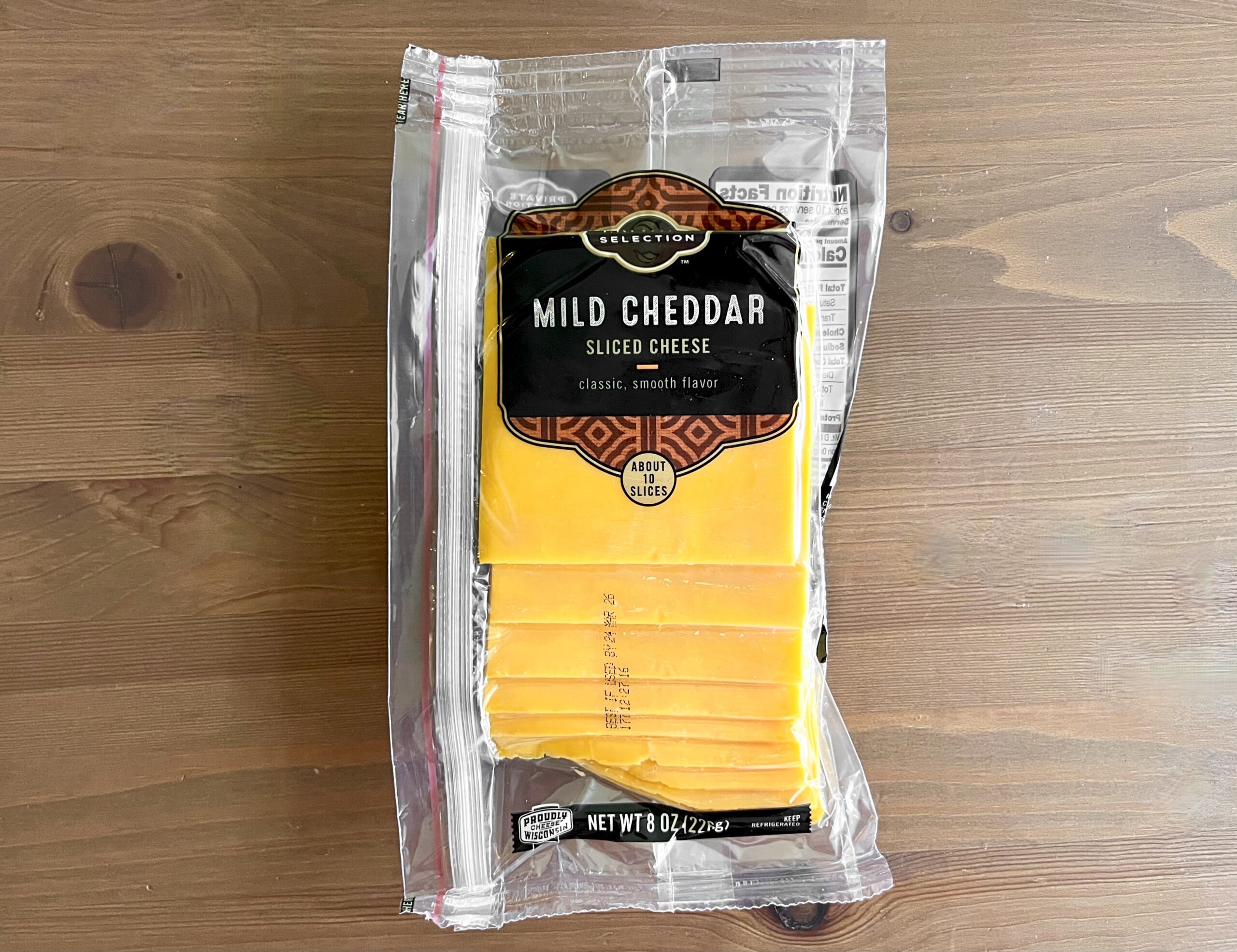 A clear plastic package of Selection brand mild cheddar sliced cheese sits on a wooden surface. The label is black and orange, and several bright yellow cheese slices are visible inside the packaging.