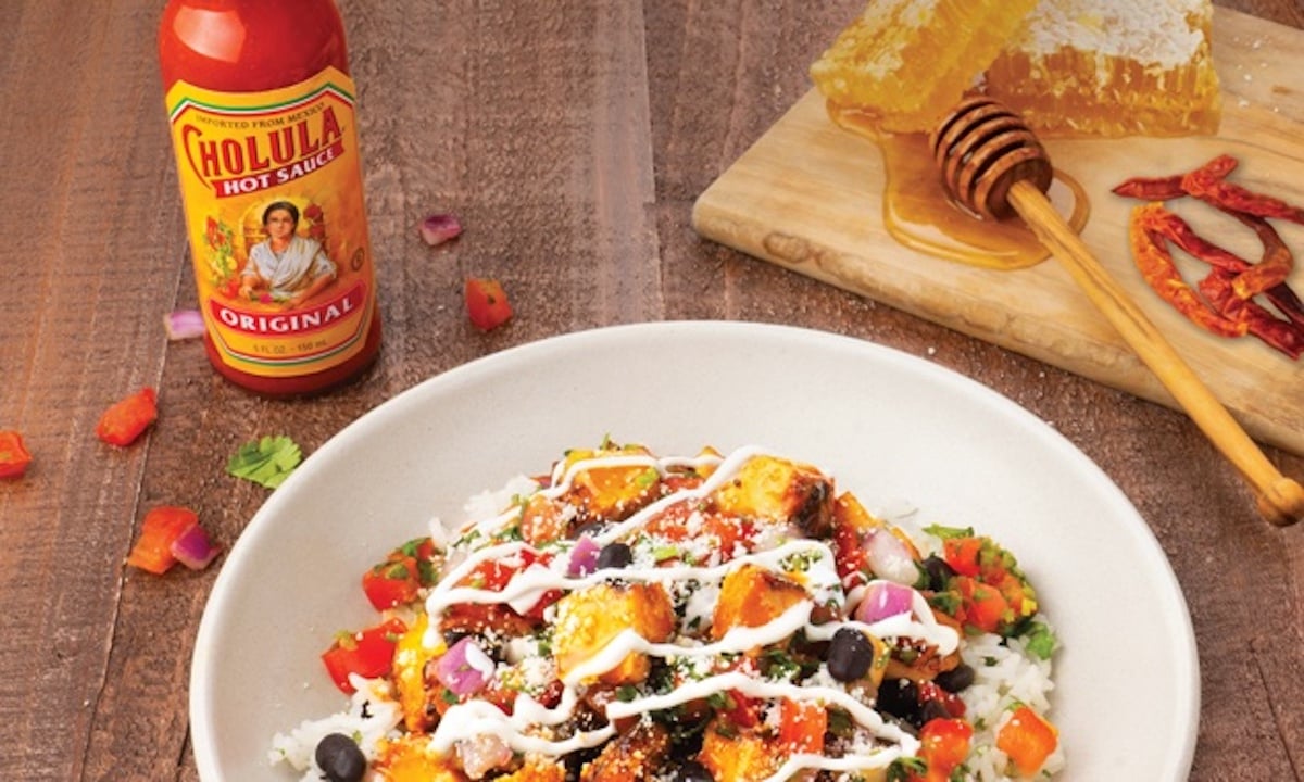 A bowl of rice topped with grilled chicken, black beans, chopped tomatoes, and a drizzle of white sauce from the new fast-food menu sits on a wooden table next to Cholula hot sauce and a board with honeycomb and dried chilis.