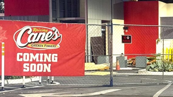 A construction site behind a chain-link fence with a large red banner reading "Raising Cane's Chicken Fingers Coming Soon." Lumber and traffic cones are visible in front of the building.