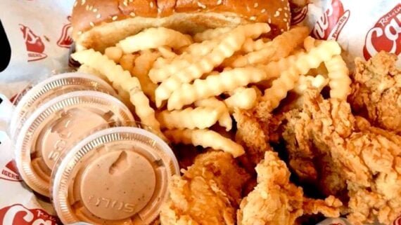 A serving of crinkle-cut fries, fried chicken tenders, three cups of dipping sauce, and a sesame seed bun on branded paper.