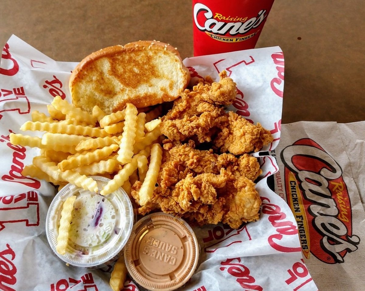 A meal from Raising Cane's featuring crinkle-cut fries, fried chicken tenders, a slice of Texas toast, coleslaw, Cane's sauce, and a soft drink in a branded cup, served on branded paper.