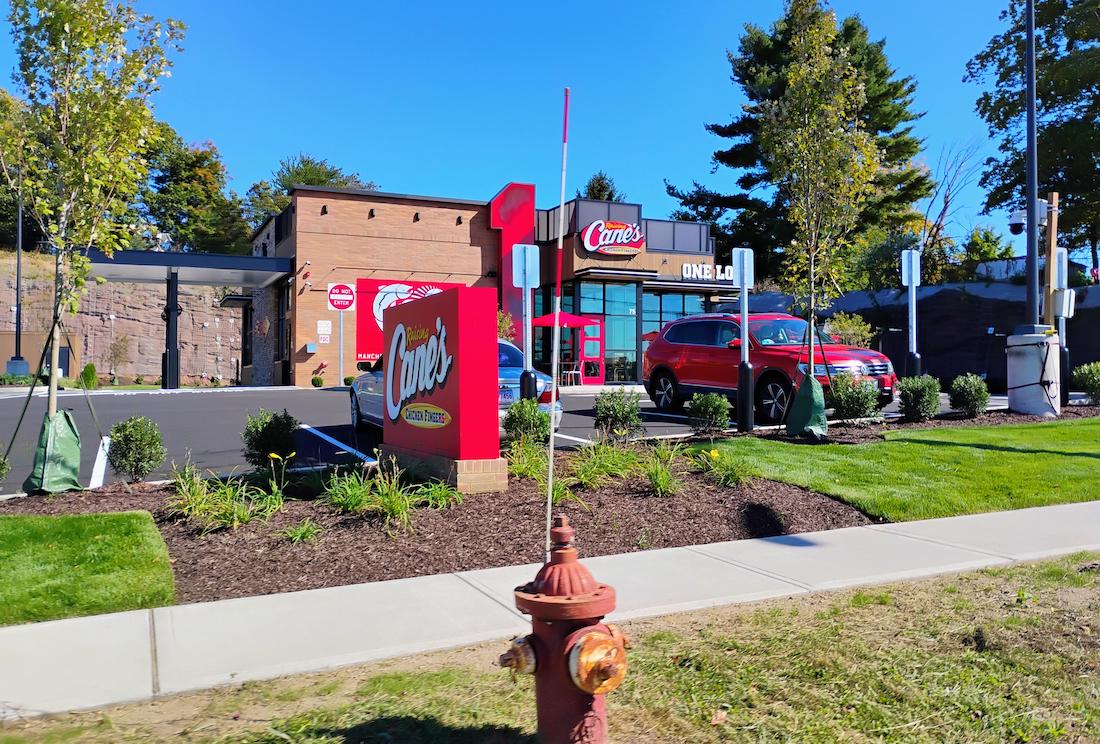 A Raising Cane's restaurant with a red sign and drive-thru, surrounded by greenery and a sidewalk. A red SUV is parked in front, and a fire hydrant is on the grass near the street.