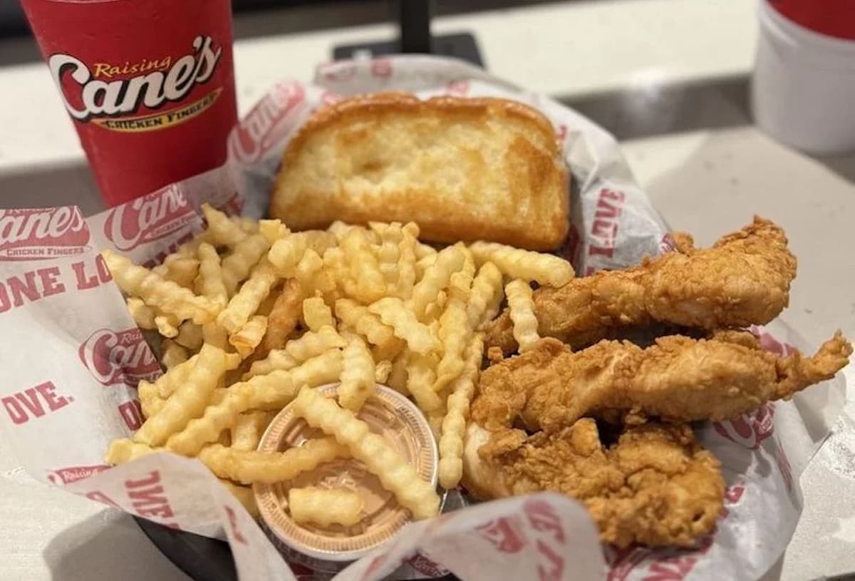 A basket of crinkle-cut fries, fried chicken tenders, toasted bread, a cup of dipping sauce, and a drink in a Raising Cane’s cup—classic fast food from one of the fast-food chains that never use a microwave, all served on branded paper.