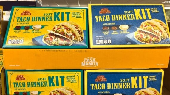 Several boxes of Casa Mamita Soft Taco Dinner Kit are stacked on a store shelf. Each box displays images of tacos and lists included items: tortillas, seasoning, and sauce. The boxes are blue and yellow.