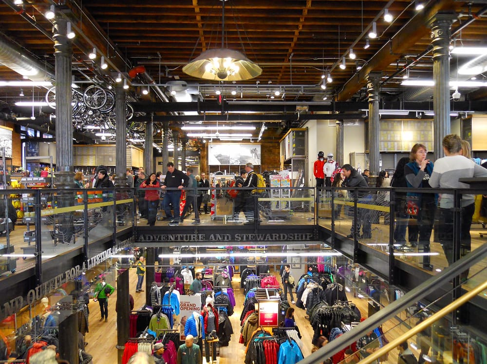 A busy outdoor gear store with two levels, filled with people shopping and talking. Colorful jackets and clothing racks are on the lower floor, while customers gather and browse on the upper level under exposed beams and hanging lights.