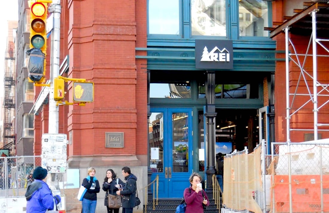 People walk past the entrance of an REI store in a red brick building, with some construction fencing and traffic lights visible on the street corner.