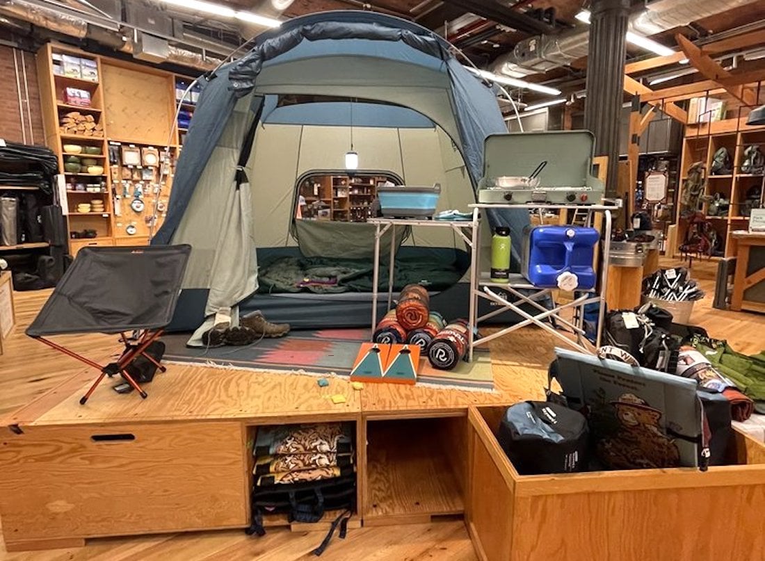 A camping setup indoors features a large tent with sleeping bags inside, surrounded by camping gear like a foldable chair, cooking stove, water bottles, blankets, and backpacks on a wooden platform in a store.