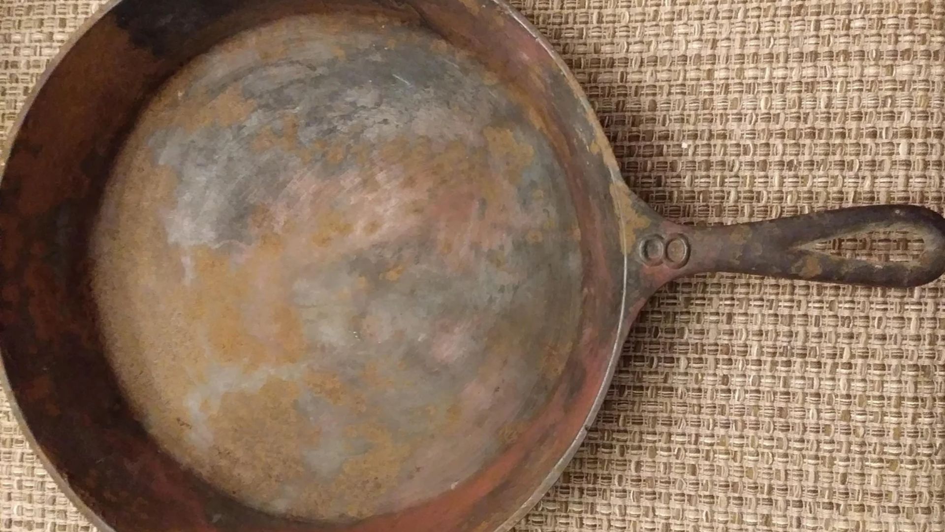A rusty, worn cast iron skillet with a handle sits on a textured burlap surface. The skillet’s surface shows discoloration and patches of rust.