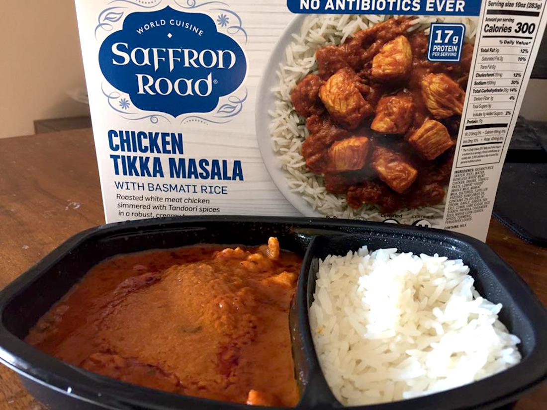 A Saffron Road Chicken Tikka Masala frozen meal is shown, with cooked chicken in orange sauce and a portion of white basmati rice in a black tray. The product box is visible in the background.