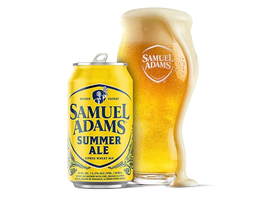 A can of Samuel Adams Summer Ale sits next to a curvy glass filled with foamy golden beer, with foam overflowing down the side of the glass. Both the can and glass display the Samuel Adams logo.