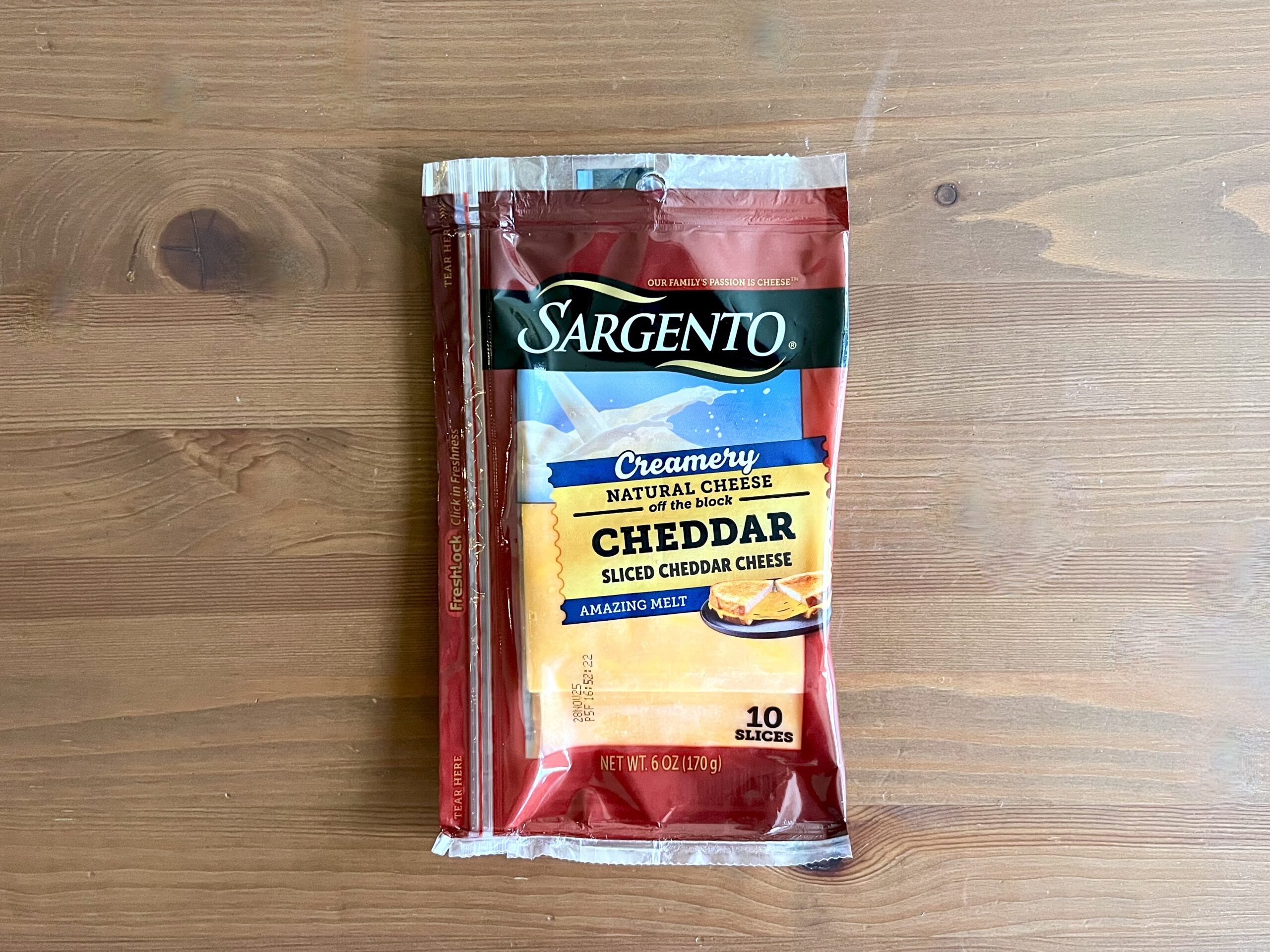 A package of Sargento Creamery Natural Cheddar Sliced Cheese with a red and yellow label sits on a wooden surface. The package contains 10 slices.