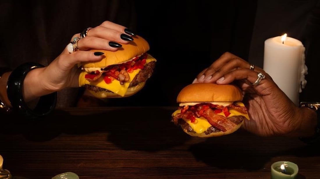 Two hands holding cheeseburgers with bacon, cheese, and toppings over a dark wooden table, surrounded by candles. One hand has dark nail polish and jewelry; the other has rings and a bracelet.