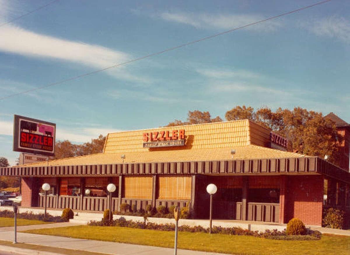 A vintage Sizzler Family Steak House restaurant with a brown brick exterior, wood accents, and a large sign in front. Bushes and globed lamps line the walkway under a clear blue sky.