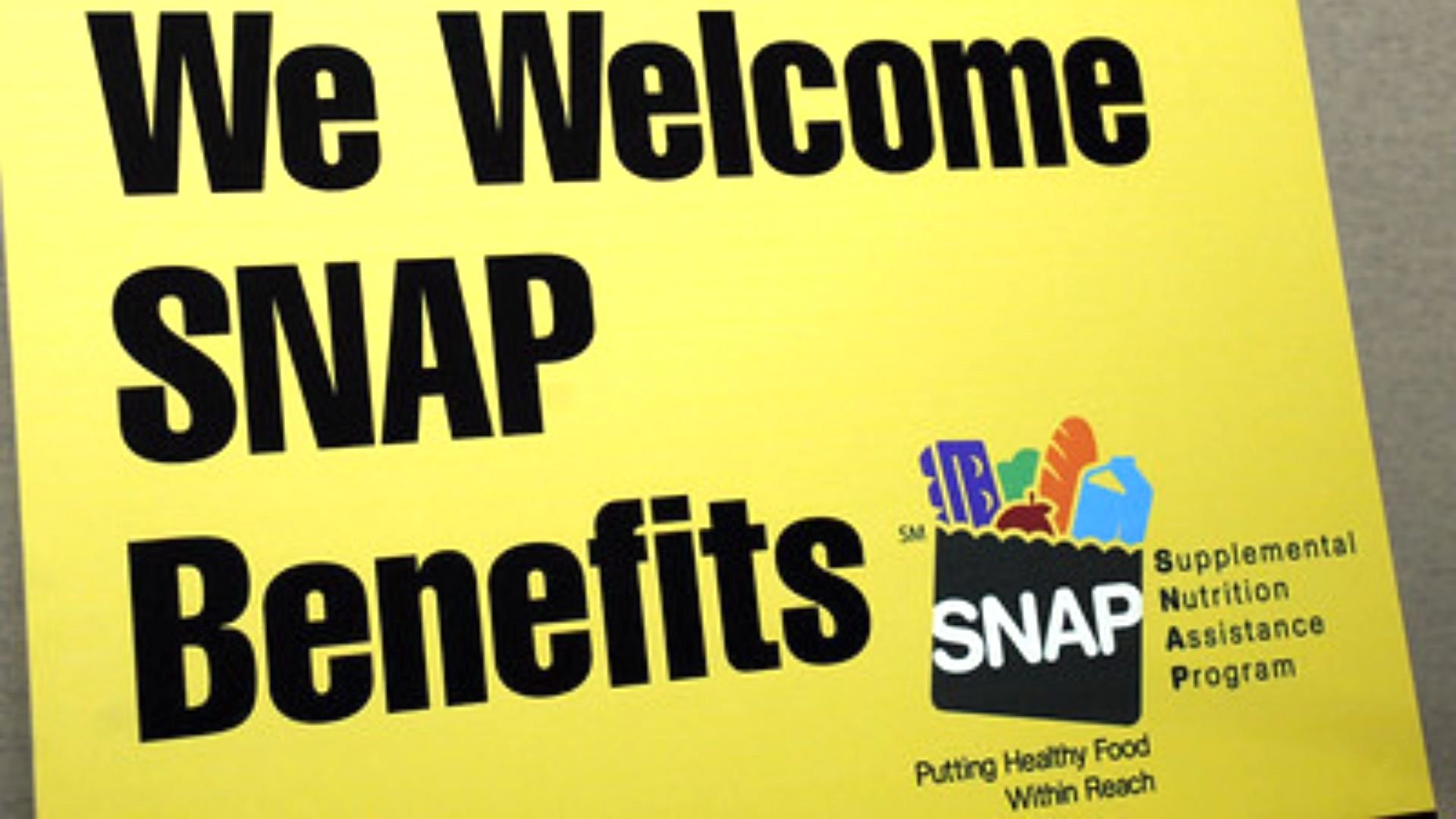 Yellow sign with bold black text reading, "We Welcome SNAP Benefits." Next to the text is a graphic of a grocery bag with food and the SNAP logo, representing the Supplemental Nutrition Assistance Program.