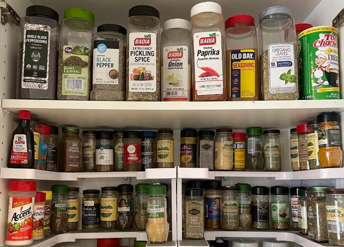 A cabinet filled with assorted spice jars and containers, organized on three shelves. Brands and labels such as black pepper, paprika, oregano, Old Bay, and others are visible, showcasing a wide variety of seasonings.