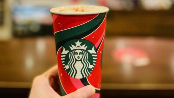 A hand holds a festive Starbucks cup with a red, green, and white holiday design and the Starbucks logo, with a blurred background of a coffee shop.
