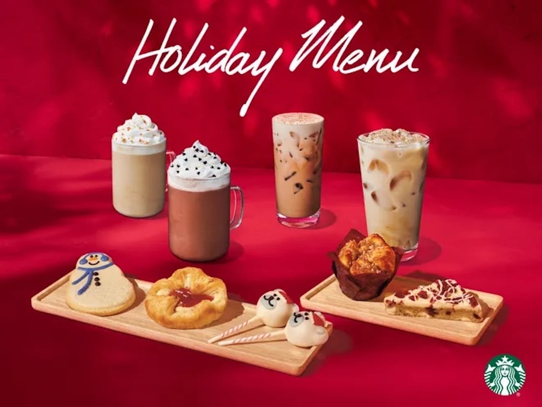 A festive Starbucks holiday menu display with four holiday-themed drinks and six assorted holiday pastries, including a snowman cookie, on wooden trays against a red background with "Holiday Menu" written at the top.