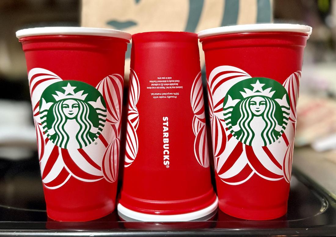 Three red Starbucks holiday cups are arranged side by side; the two on the sides are upright, while the middle one is upside down. All cups display the Starbucks logo with festive white and green designs.