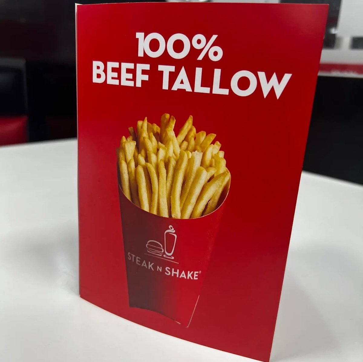 A red table sign displays an image of French fries in a red container with the Steak 'n Shake logo, above bold white text that reads “100% BEEF TALLOW,” highlighting an exciting addition to the new fast-food menu.