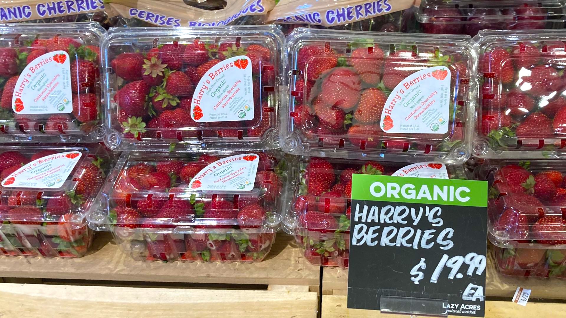 Plastic containers filled with organic strawberries labeled "Harry's Berries" are stacked on a wooden display. A green and black sign reads "ORGANIC HARRY'S BERRIES $19.99.