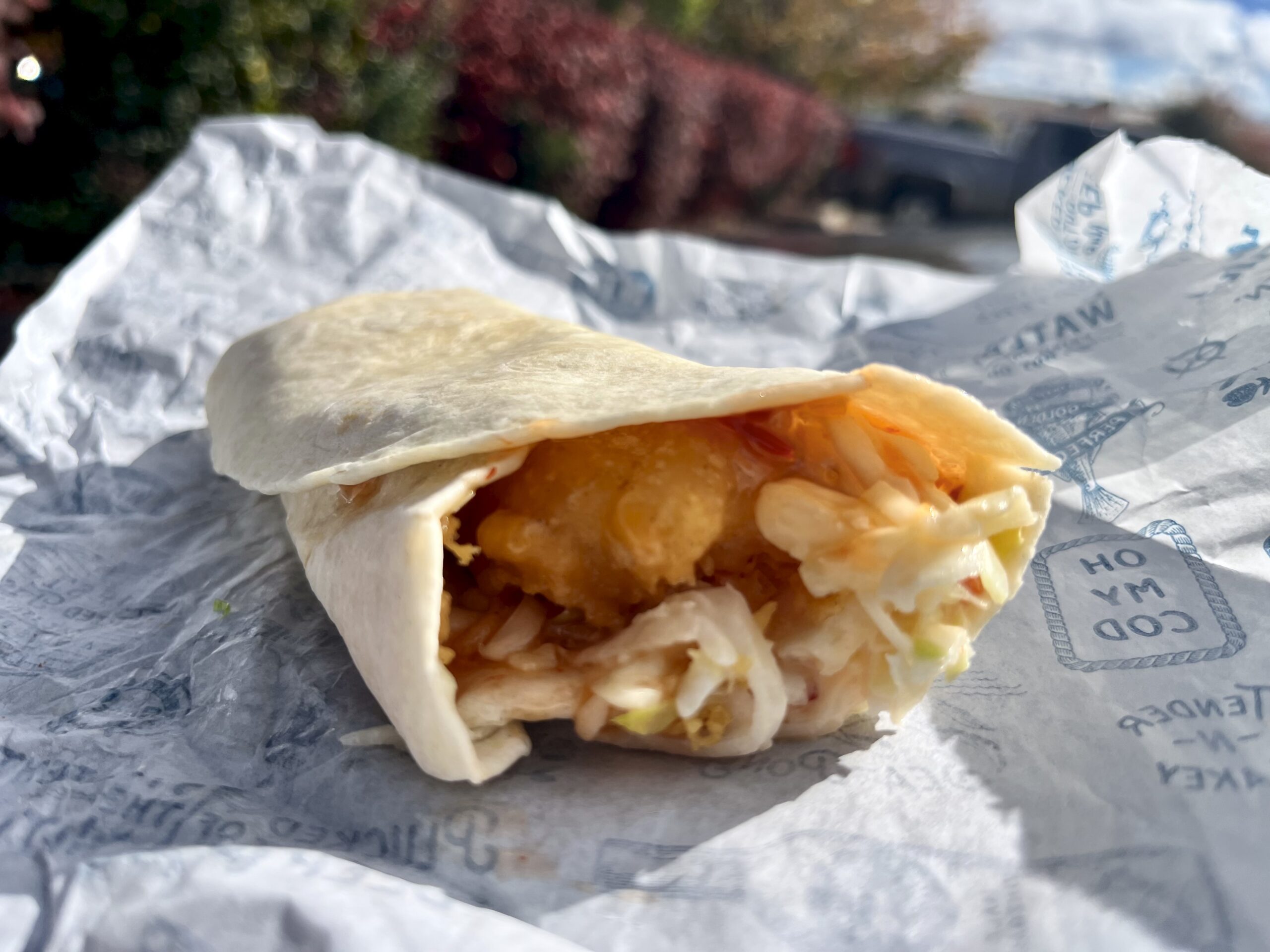 A close-up of a partially unwrapped tortilla wrap filled with fried fish and shredded cabbage, resting on crumpled paper outdoors with blurred trees and a truck in the background.