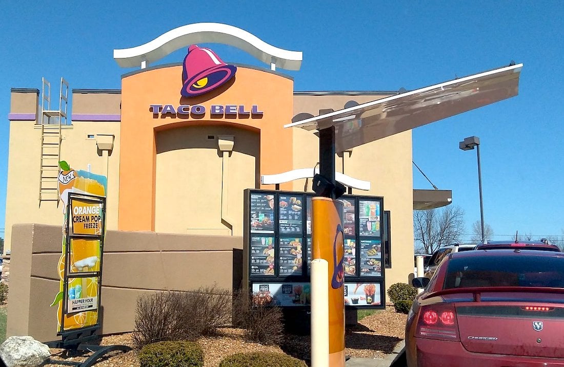 Cars line up at a Taco Bell drive-thru on a sunny day, with a menu board and signs visible near the restaurant’s tan building and purple bell logo. Shrubs and clear blue sky are in the background.