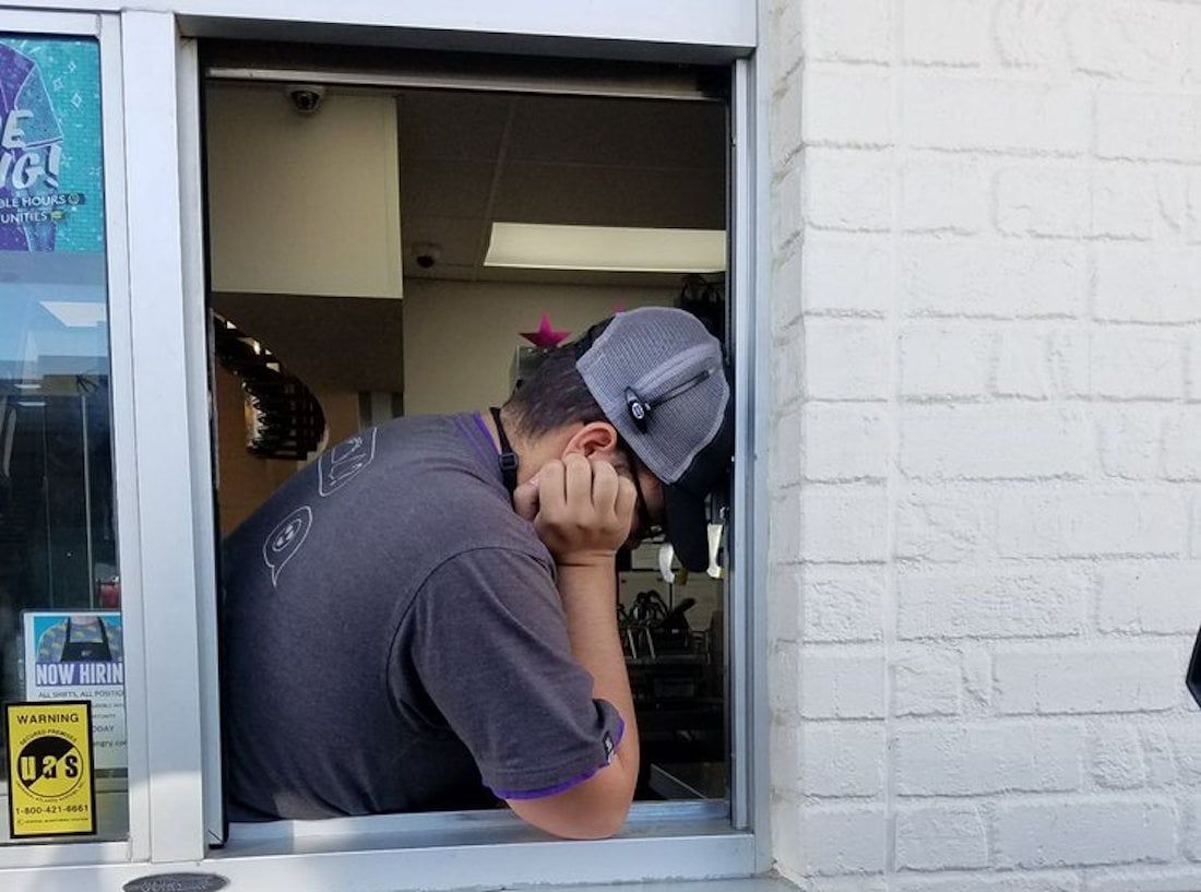 A fast-food worker in uniform leans on the drive-thru window with his head resting on his hand, appearing tired or stressed. Signs and stickers are visible on the window beside him.