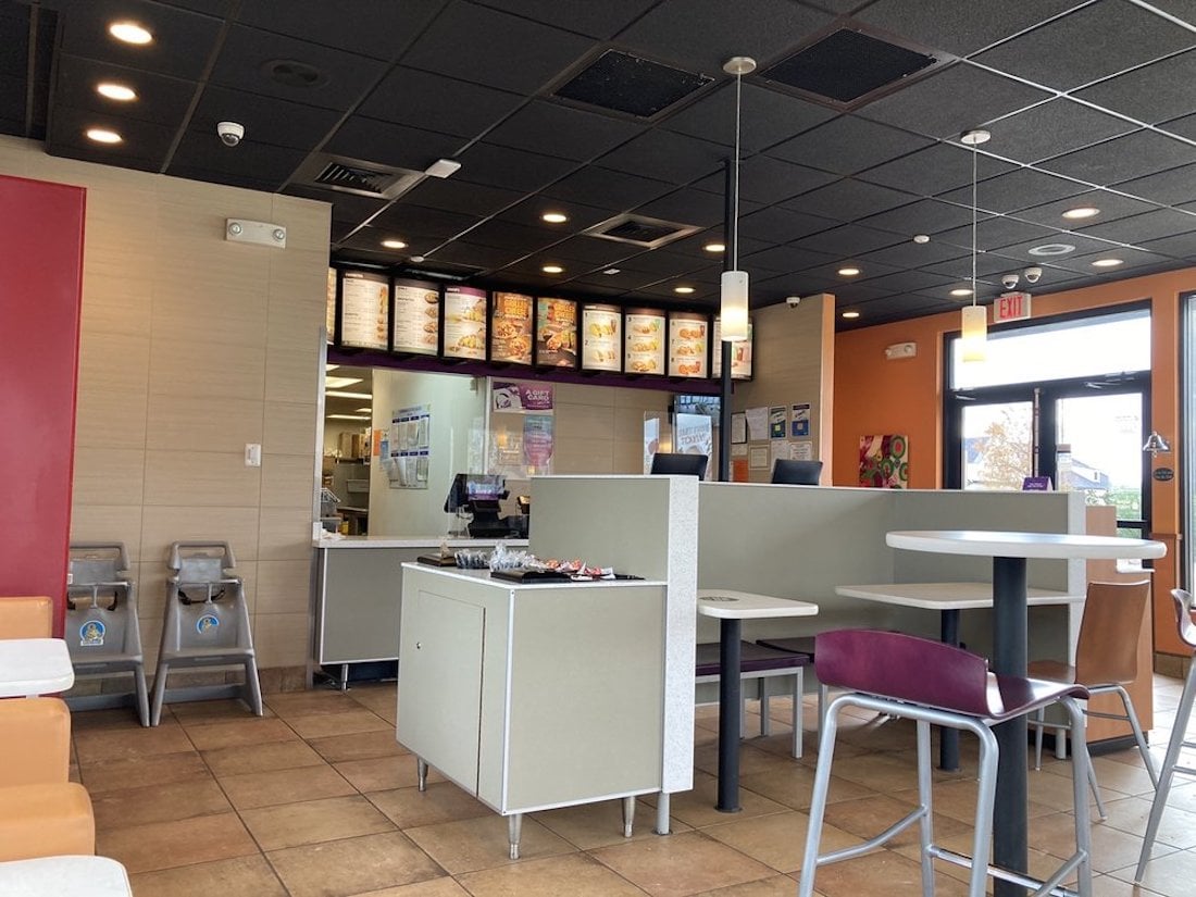 Interior of a fast-food restaurant with empty tables and chairs, an ordering counter with menu boards above, condiment trays on a counter, and highchairs against the wall. The space is brightly lit with tile flooring.