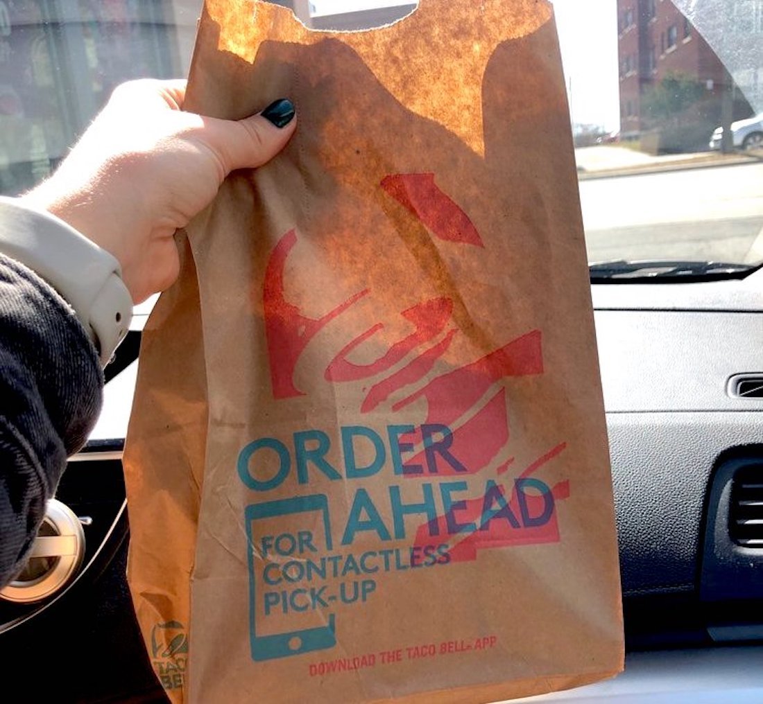 A hand holding a Taco Bell paper bag inside a car. The bag displays text promoting ordering ahead for contactless pick-up. Sunlight shines through the car window, and buildings are visible outside.