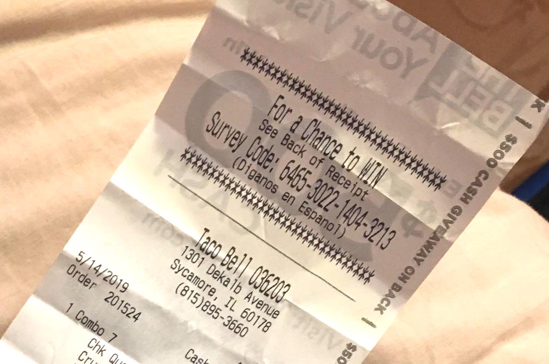 A close-up of a Taco Bell receipt showing a survey code, date, time, order details, and the restaurant's address in Sycamore, Illinois. The text is slightly tilted and partially cropped at the bottom.
