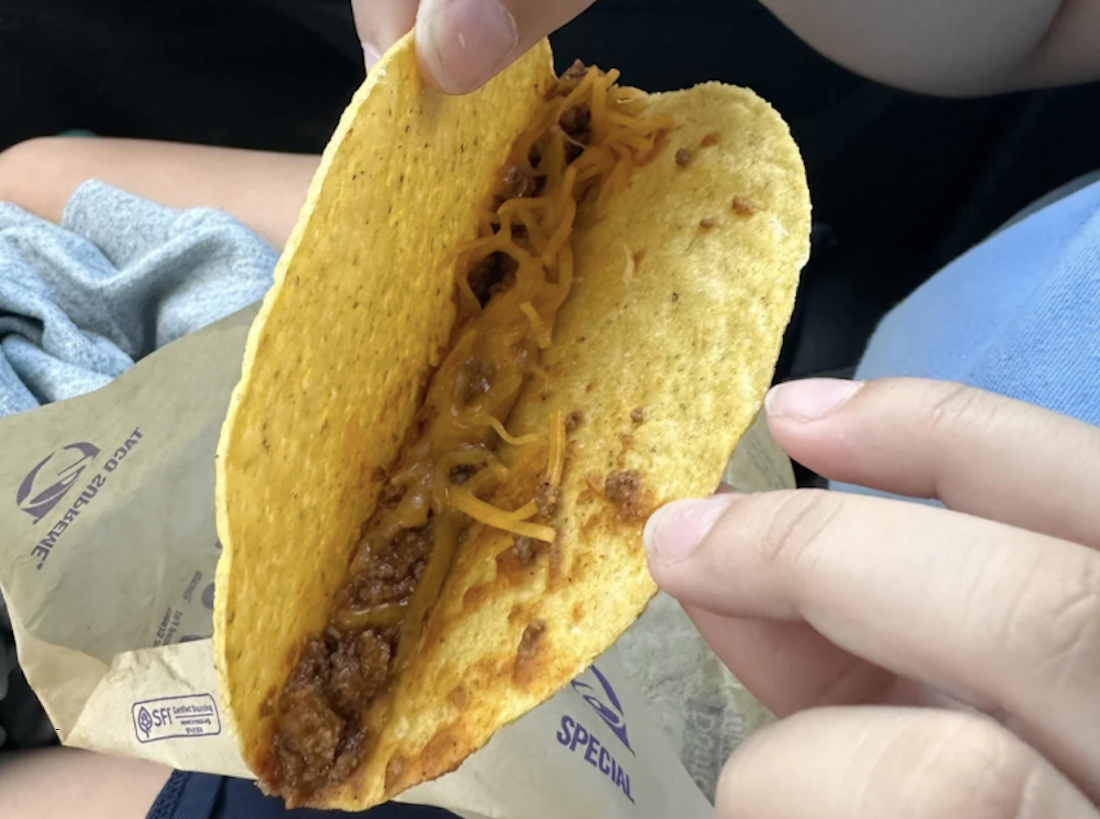 A person holds a hard taco shell filled with ground beef and shredded cheddar cheese over a Taco Bell wrapper, with hands and legs visible in the background.