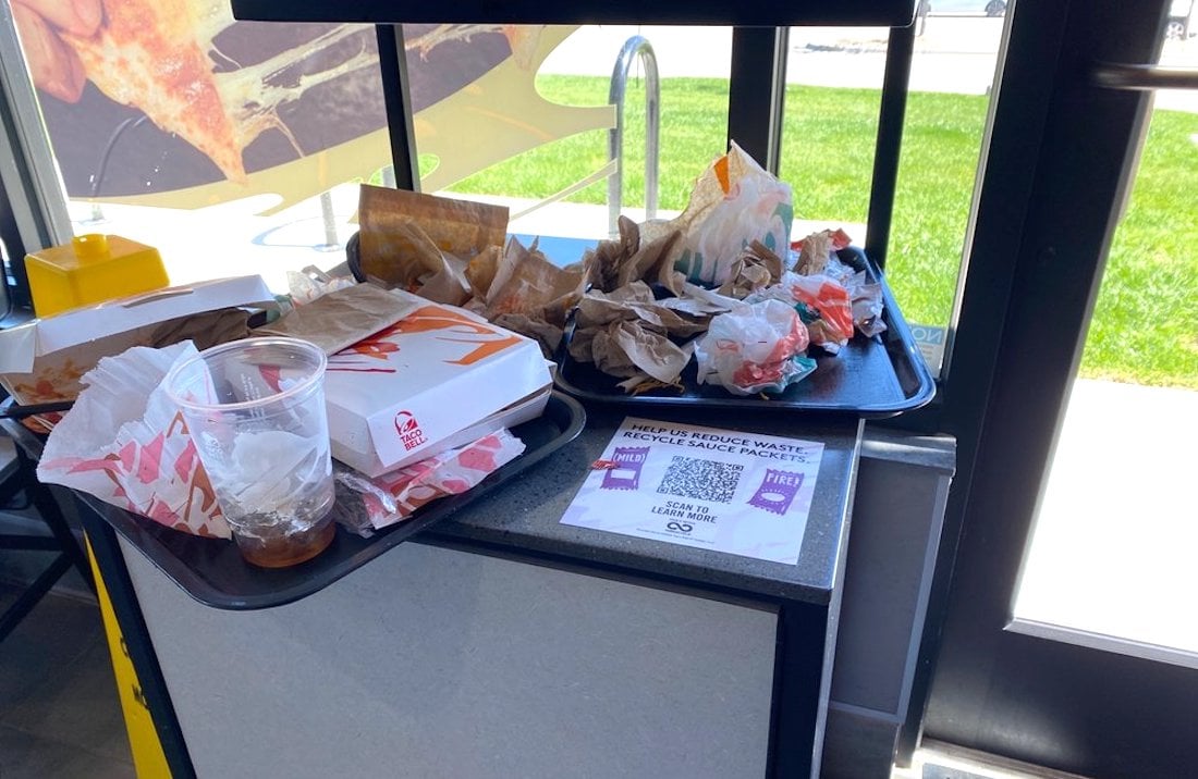 A trash station in a fast food restaurant is overflowing with trays, empty cups, food wrappers, napkins, and other discarded items, with a bright window and green grass visible outside.