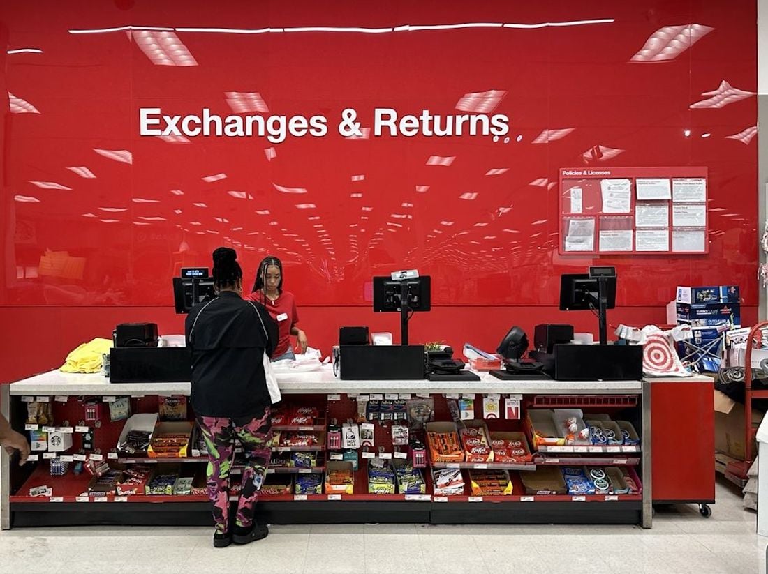 A customer stands at a store's "Exchanges & Returns" counter, speaking with a staff member. The background is a bright red wall with white text and posted notices. Various items and snacks are displayed on the counter.
