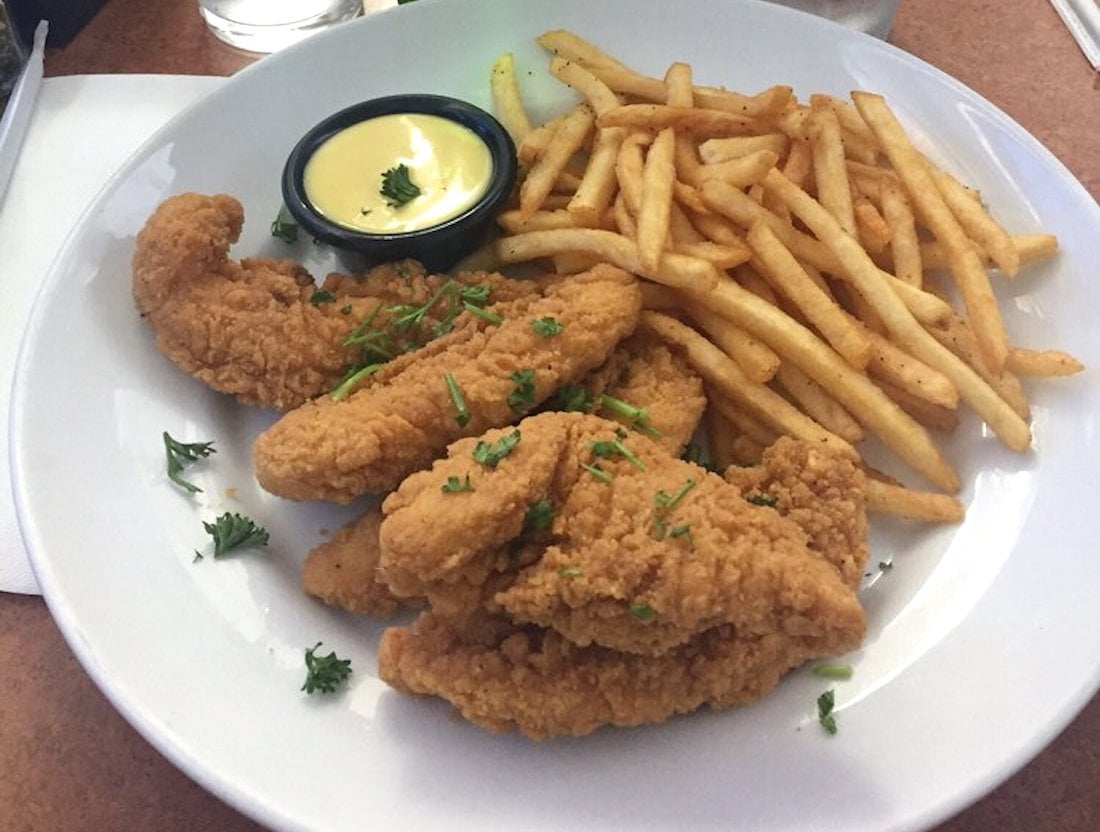 A white plate with crispy fried chicken tenders, a serving of French fries, a small cup of yellow dipping sauce, and a garnish of chopped herbs.